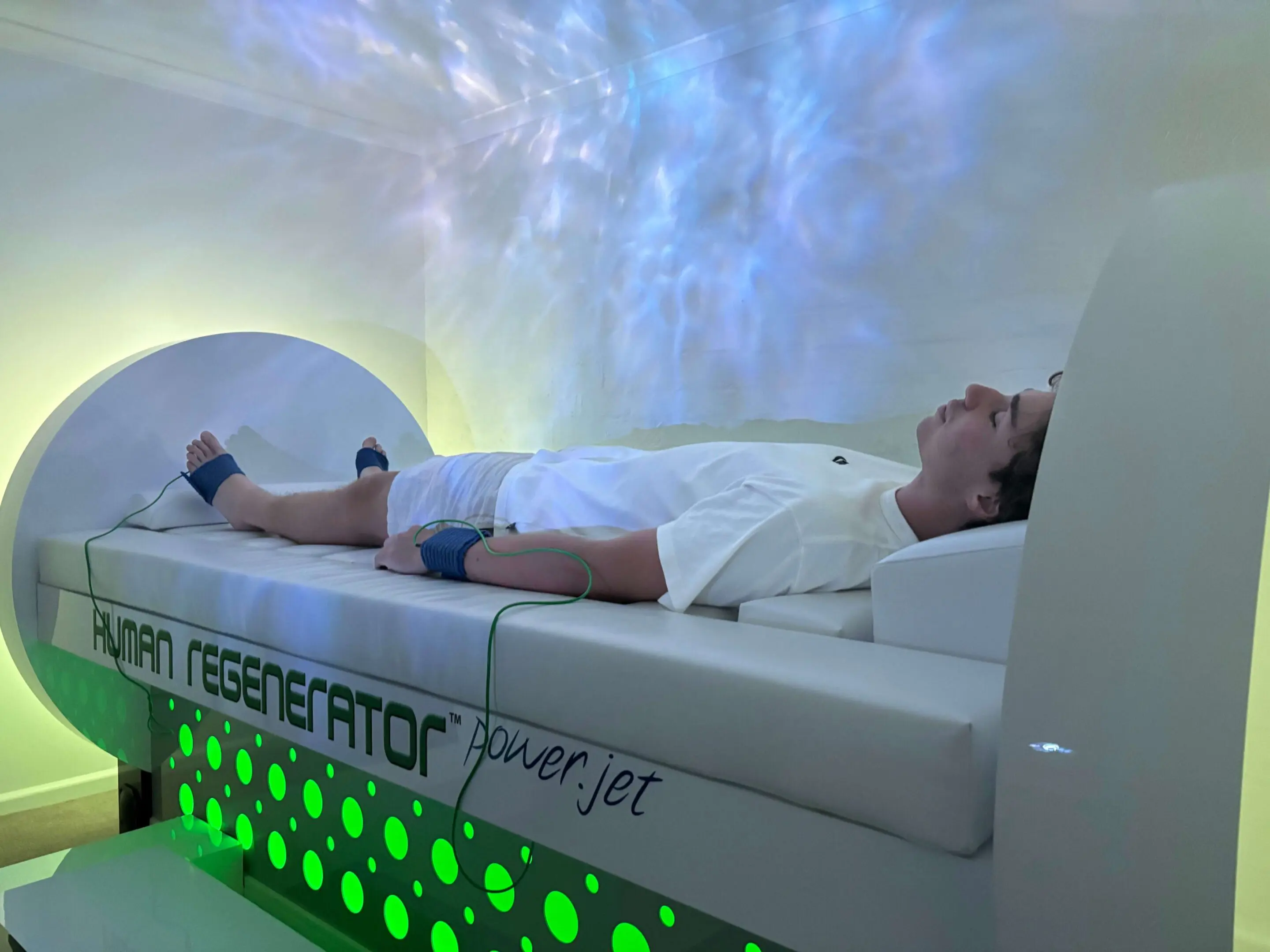 IMG_9984 A person lies on a futuristic bed labeled "Human Regenerator," surrounded by glowing green lights. Dressed in white and connected to wires, they experience the bioenergy healing benefits of this advanced technology.