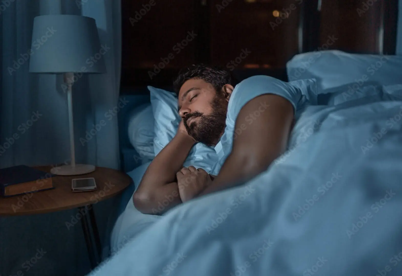 A man sleeps peacefully in bed at night, embraced by the calming aura of bioenergy healing. Beside him, a lamp casts a gentle glow over his phone on the bedside table, enhancing his restorative rest.